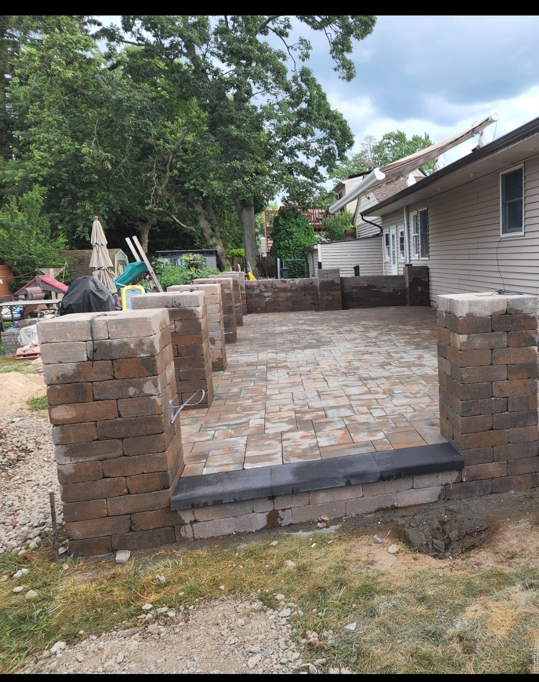 A partially constructed patio with stone pavers and brick pillars by a house showcases the work of a skilled home remodeler in Suffolk County, NY. Dirt, grass, trees, and outdoor furniture surround the inviting space in progress.