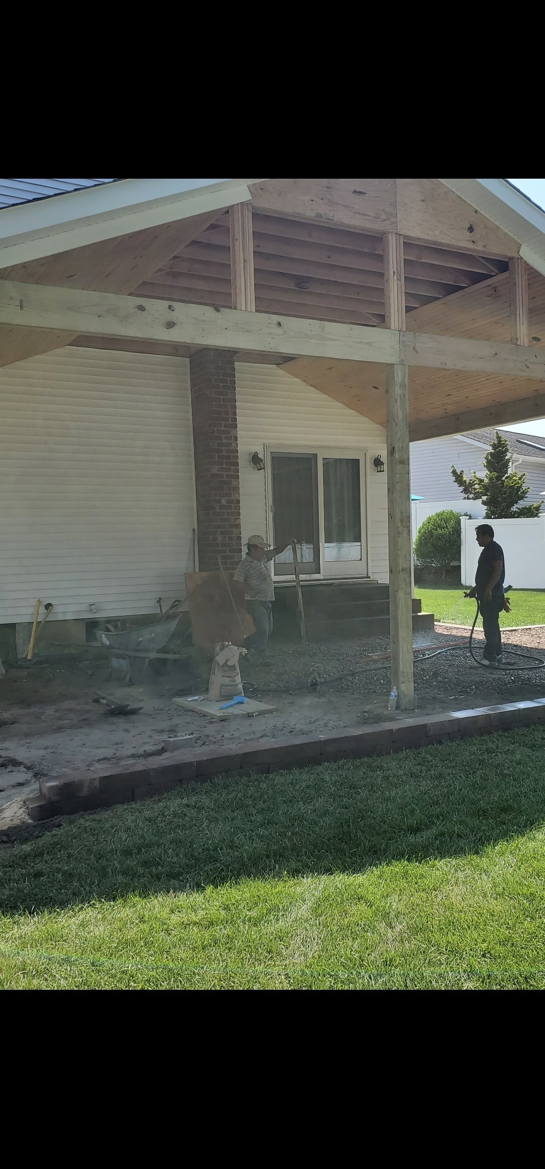 Two people work under a partially constructed wooden patio roof attached to a house. One, possibly a home remodeler in Suffolk County, NY, uses a shovel while the other stands nearby amid scattered materials and grassy foreground.
